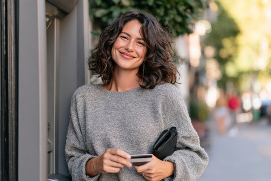 Smiling curly haired woman holds a payment card beside an outdoor ATM. Personal banking, card access, financial confidence, urban lifestyle.