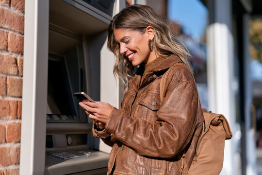 Smiling young woman in a brown leather jacket checks her phone at an ATM. Mobile banking, cash access, urban lifestyle, personal finance.