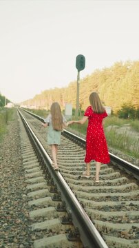 Two young women hold hands as they stroll down parallel railroad tracks. One wears striped shirt with light green overalls other is dressed in red dress with white patterns. scene features