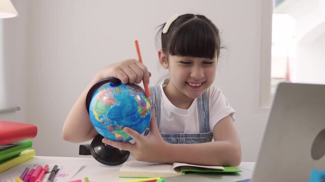 Cute Asian schoolgirl sitting at a desk using laptop computer for studying online learning via video calling and a remote teacher while having a geography lesson with a globe model in homeschool room.