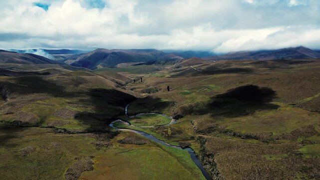 A dark, winding stream crosses open highland grassland, bordered by pale gravel and sand bars and patches of green and brown vegetation. Aerial drone footage captures the broad, textured landscape of 