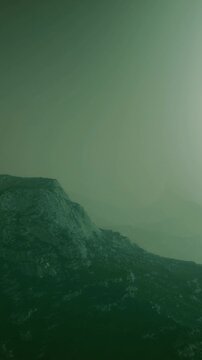 Fog covers the mountain landscape during early morning hours. The scene shows various shades of green and gray. The time appears to be at dawn with soft light touching the peaks.