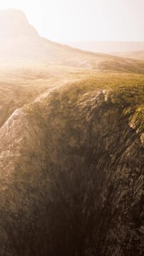 A large rocky cliff is visible under bright sunlight in a mountainous area. The ground is mostly dry and has patches of green grass. The scene looks open and vast.