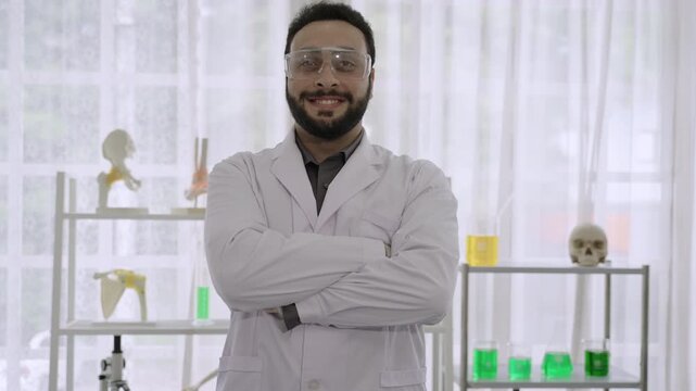 Portrait of happy handsome Middle Eastern young man scientist wearing a white lab coat and safety goggles, smiling and standing with arms crossed while looking at the camera in a science laboratory. 