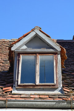 An old weathered wooden dormer window with peeling paint is built into a traditional clay tiled house roof under a blue sky.