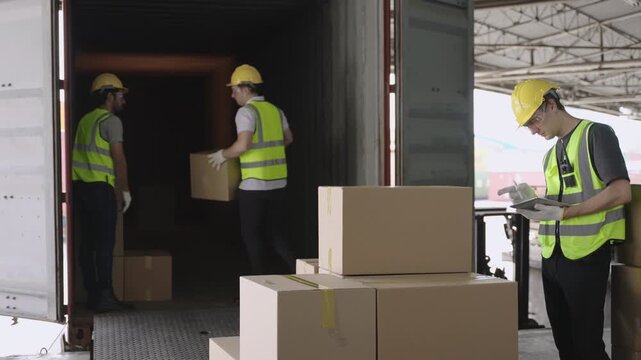Team of young men warehouse workers in uniform wearing yellow safety helmets helping, holding, and carrying cargo boxes for loading into a container before delivery in a logistics distribution factory