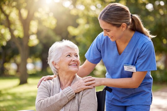 A compassionate caregiver in blue scrubs assists and comforts a smiling elderly woman in a wheelchair outdoors in a sunny park.