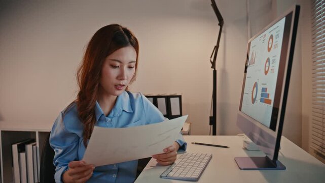 Focused young asian businesswoman comparing financial reports on paper with charts and graphs on her computer screen while working late in the office to meet a project deadline