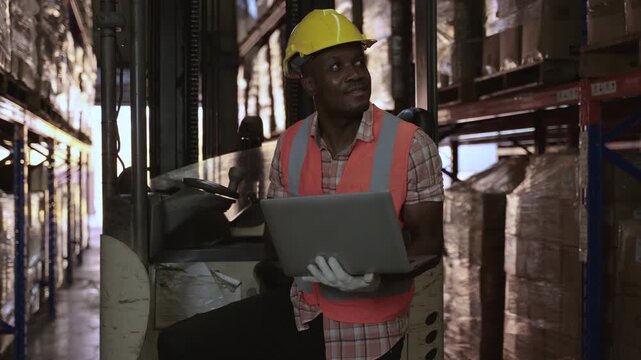 Professional African American male forklift driver wearing orange vest and yellow safety helmet using a laptop to inventory stock on shelves before delivery in logistics distribution warehouse center.