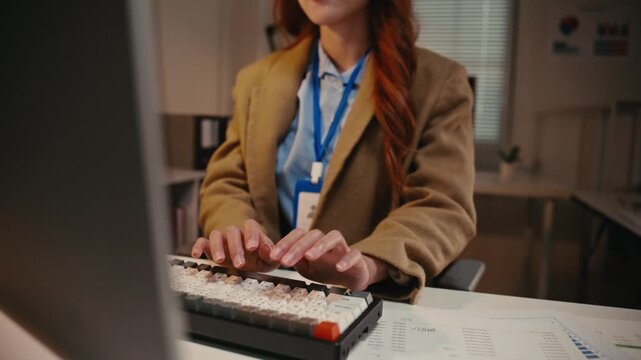 Young Asian business professional diligently typing on a computer keyboard, analyzing financial charts and data while working overtime in a modern corporate office during the evening hours
