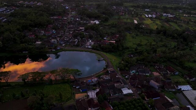 Aerial view of Danau Batu Karut reflecting the sunset sky, surrounded by lush village greenery and residential rooftops in Sukabumi, West Java, Indonesia.