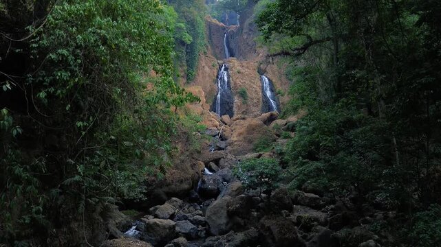 Aerial drone footage of a scenic waterfall flowing over rocky terrain through a dense tropical forest. Multiple cascading streams descend between mossy rocks and lush green vegetation.