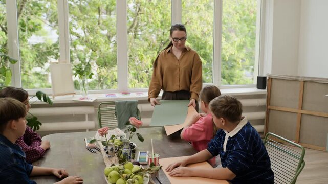Caucasian female teacher distributing colored paper sheets to children during creative art master class. Perfect for educational marketing and childhood development projects