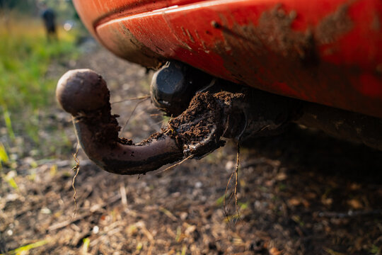 A vehicle tow ball and curved towbar carry clumps of mud beneath the rear bumper beside a trailer socket, showing towing hardware relevant to cleaning and hitch maintenance