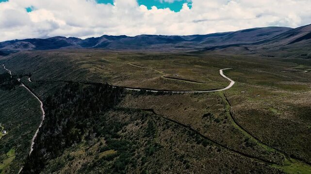A wide aerial view frames a dark blue lake in the foreground, bordered by rounded Andean hills and distant mountain ridges. Thick white clouds fill much of the sky above this highland landscape, with 