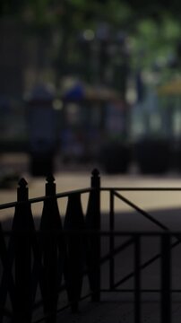 A black metal railing is in the foreground of a park scene. People walk in the background near trees and structures. The setting shows vibrant activity during daytime.