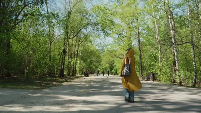 Woman wearing long bright yellow garment and carrying patterned bag strolls along sunlit park pathway. Distant figures walk or sit among trees under clear blue sky