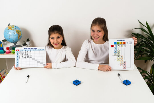 Two happy girls proudly displaying completed mathematics learning boards with a multiplication table and long division exercise while studying together.