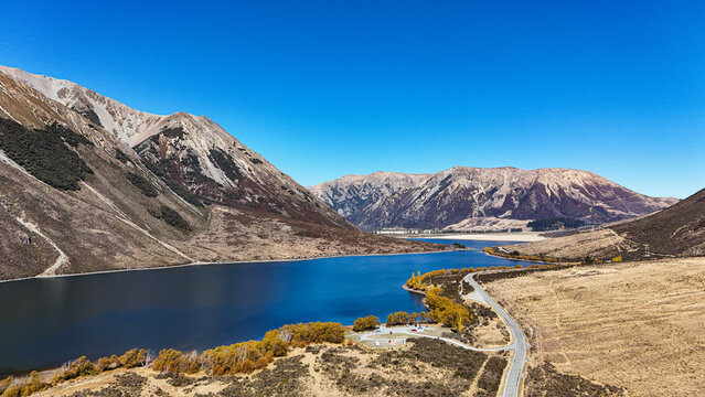 Aerial view of Lake Pearson in Arthur's Pass, featuring blue water, rugged mountains, golden autumn trees, and a winding road, Arthur's Pass, Canterbury Region, New Zealand.
