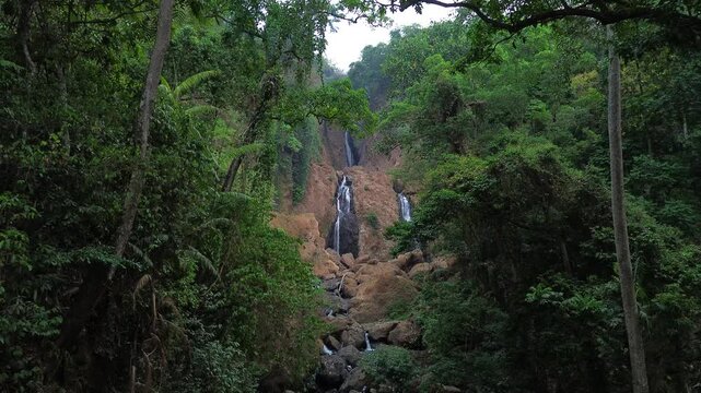 Aerial drone footage of a scenic waterfall flowing over rocky terrain through a dense tropical forest. Multiple cascading streams descend between mossy rocks and lush green vegetation.