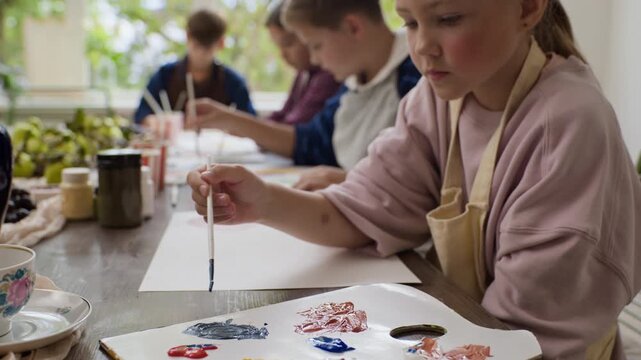 Caucasian girl dipping brush in paint palette and painting during creative masterclass with children. Ideal for art school advertising and education workshop marketing