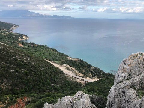 Stunning high-angle panoramic view from a rocky mountain peak overlooking a winding asphalt road, lush green slopes, and calm turquoise sea under a cloudy sky in Greece