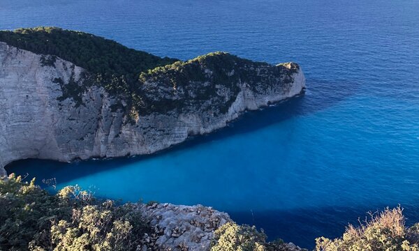 A high-angle view of the dramatic limestone cliffs and the vibrant, glowing turquoise water of Navagio Beach (Shipwreck Cove) on Zakynthos Island, Greece