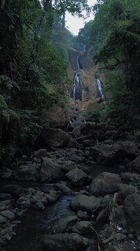 Aerial drone footage of a scenic waterfall flowing over rocky terrain through a dense tropical forest. Multiple cascading streams descend between mossy rocks and lush green vegetation.