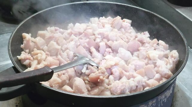 stir frying diced pork fat belly meat in black frying pan with ladle ladle spatula steam smoke rising up close up angle
