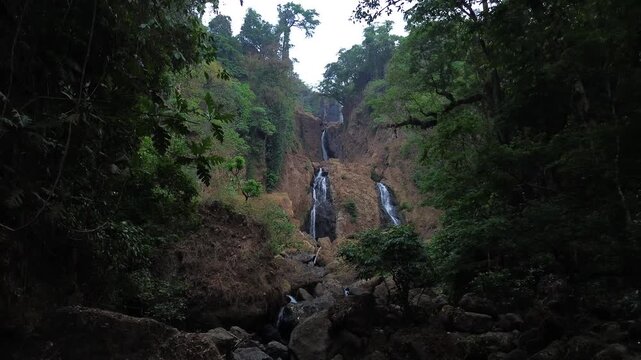 Aerial drone footage of a scenic waterfall flowing over rocky terrain through a dense tropical forest. Multiple cascading streams descend between mossy rocks and lush green vegetation.