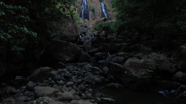 Aerial drone footage of a scenic waterfall flowing over rocky terrain through a dense tropical forest. Multiple cascading streams descend between mossy rocks and lush green vegetation.