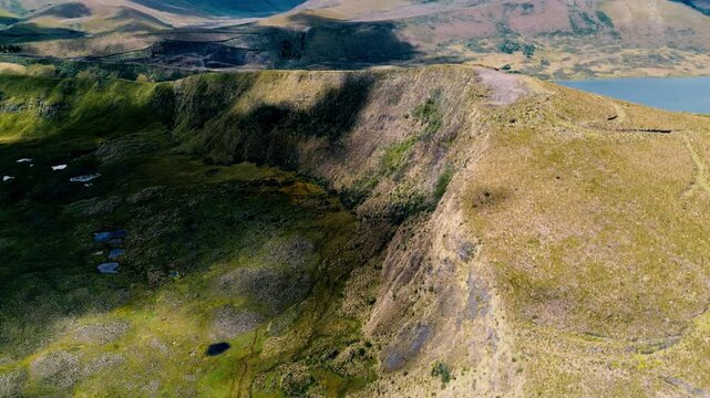 Aerial footage over the Andes in Ecuador reveals a broad grassy ridge and steep escarpment above scattered wetland pools. A blue lake, rolling mountain slopes and cloud shadows complete the highland l