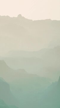 Fog covers the mountains as the sun rises, creating a soft light. The hills and valleys show a mix of grey and blue colors, creating a mysterious scene at dawn.