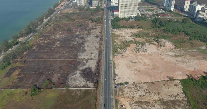 Tilting up aerial view reveals the stark contrast between the sprawling Sihanoukville city skyline and the vast, undeveloped land plots separated by a long road