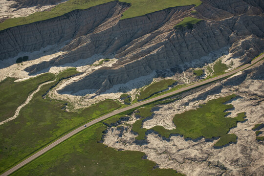 Aerial view of Badlands National Park with eroded rock formations, rugged terrain, green grassy patches, and a winding road cutting through the landscape in Interior, South Dakota, United States.