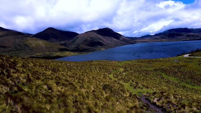 A steep, sparsely vegetated escarpment borders a broad highland hollow dotted with small ponds, surrounded by green and tawny grassland. This aerial view of the Andes in Ecuador highlights the rugged 