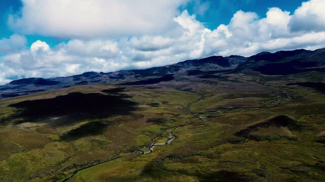 A drone aerial view looks across the open Andean highlands of Ecuador, where rolling green and brown terrain leads toward rugged mountain ridges. A narrow road curves through the foreground, and a win