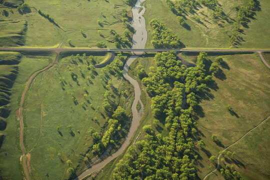 Aerial view of Badlands National Park featuring a winding river, lush green trees, and a bridge crossing the landscape in Interior, South Dakota, United States.