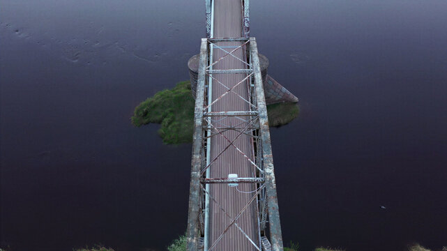 Aerial view of a metal truss bridge spanning a dark river with a small grassy island in Latvia.