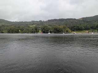 Scenic calm lake landscape view with green forested hills, serene water surface, and sailing boats anchored near the shore © NUMUD