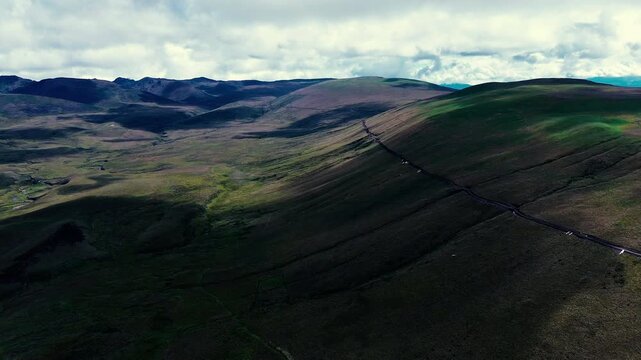 A drone aerial view looks across rolling green and brown ridges in the Andes of Ecuador, where a narrow winding road crosses the highland terrain. Layered mountain ranges recede beneath heavy clouds, 
