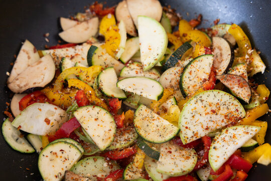 Sliced zucchini and bell peppers cooking in a pan with spices against a dark metal background