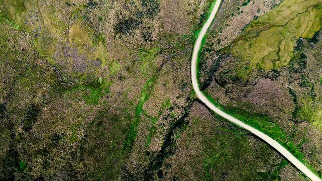 Drone footage surveys a narrow road running diagonally across broad, rounded hills in the Andes of Ecuador. Green and brown highland slopes extend toward layered mountain ridges beneath a cloud-filled