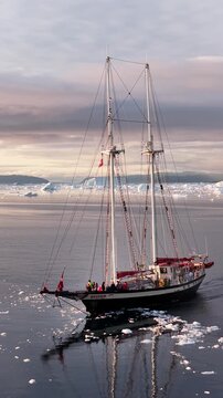 Aerial view of a two-masted schooner navigating through the icy waters of the Ilulissat Icefjord with icebergs in the background in Ilulissat, Avannaata Municipalit, Greenland.