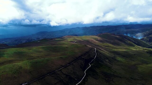 An aerial view across the Andes of Ecuador reveals green and brown uplands, layered mountain ridges and a narrow stream winding through a broad valley. Broken cloud cover casts shadows across the terr