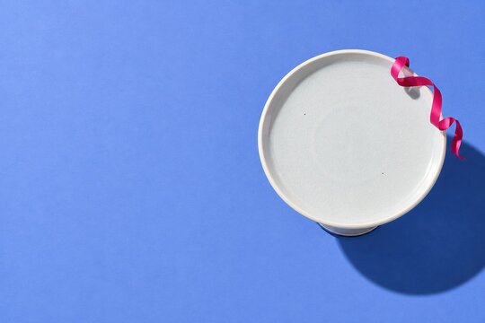 Empty white ceramic cake stand on a periwinkle blue background, a clean blank mockup with a fuchsia ribbon for holiday displays