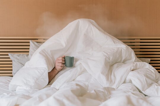 Man hidden under a thick white blanket in bed holding a steaming coffee cup, cozy and humorous scene for winter cold and sick day
