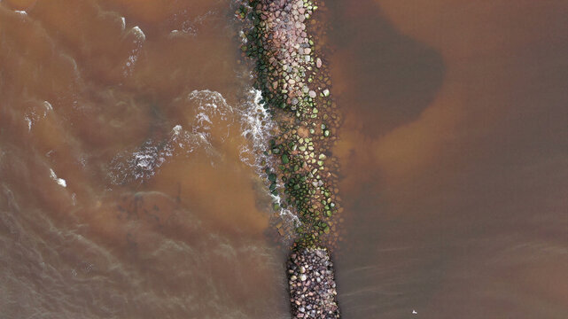 Aerial view of a stone breakwater extending into the sea, with waves crashing against the moss-covered rocks in Latvia.