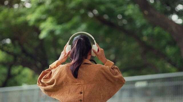 Rear view of young Asian woman putting on wireless headphones outdoors in green urban park, adjusting headset before listening to music, enjoying nature, digital audio technology.