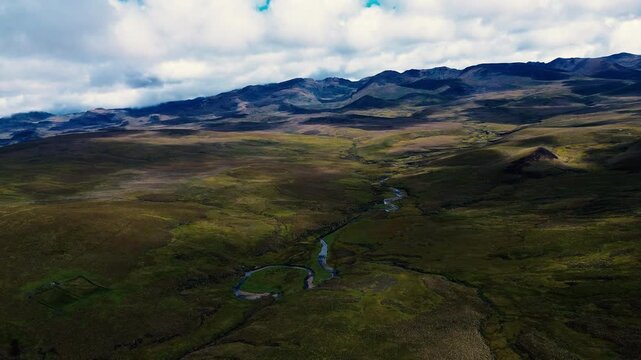 Drone footage provides an oblique aerial view of a pale winding dirt road crossing open, rolling highland grassland in the Andes of Ecuador. A narrow stream and bands of greener vegetation cut through